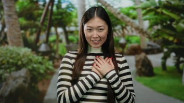 Kadın açık hava parkında duruyor, elleri göğsünde, çizgili tişört giyiyor, yemyeşil, sıcacık gülümsüyor, huzurlu, huzurlu bir atmosfer yaratıyor..