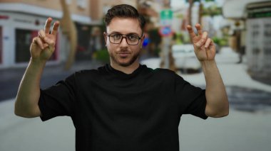 Sakallı genç bir adam, şehir merkezinde gün ışığında dışarıda dikilirken gözlük ve günlük giysiler gösteren bir sokakta barış işareti yapıyor..