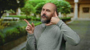 Şehir sokağında yeşil arka planda, kel, sakallı, gri kazak giyen, kendine güvenen, yakışıklı bir yüz ifadesi olan, açık hava manzaralı, şehir manzaralı bir adam..