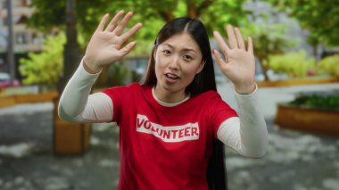 Gönüllü gömleği giyen, kolları parkta yükselen, arka planda ağaçlar ve binalar olan genç bir kadın, coşku ve açık havada toplum ruhunu gösteriyor..