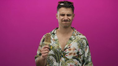 Genç İspanyol adam canlı pembe arka planda dondurmadan zevk alıyor duygularını ifade ediyor, başına güneş gözlüğü takıyor, kaygısız bir turist havasını somutlaştırıyor..