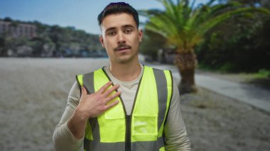 Yansıtıcı yeleği olan genç bir adam parkta dikiliyor. Palmiye ağaçlarının ve açık gökyüzünün önünde düşünceli bir ifade sergiliyor. Açık hava güvenliğini simgeliyor..