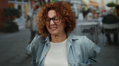 Woman experiencing back pain on a bustling city street, showcasing curly brunette hair, glasses, and a casual outfit, surrounded by urban life and outdoor seating.