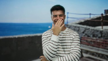 Genç adam, güneşli bir günde okyanus geçmişiyle deniz kenarında gülümsüyor.