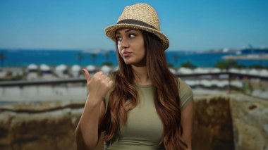 Hasır şapkalı bir kadın açık gökyüzü ile deniz manzaralı bir limana başparmağını doğrultuyor güneşli bir günde arka planda teknelerle gezen genç İspanyol havasını yansıtıyor..