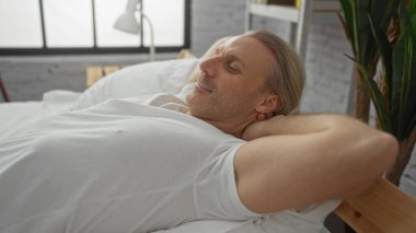 Orta yaşlı bir adam modern bir yatak odasında doğal ışıkla dinleniyor, beyaz bir gömlek giyerek rahat bir şekilde yatakta uzanıyor, huzurlu ve huzurlu bir ev atmosferi yaratıyor..