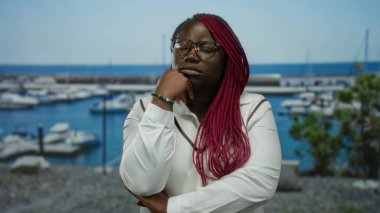 Gözlüklü ve kırmızı örgülü bir kadın, arka planda tekne ve deniz olan bir limanın yanında düşünceli bir şekilde oturur, berrak mavi gökyüzünün altında derin bir düşünce ve dinginlik hissi yayar..