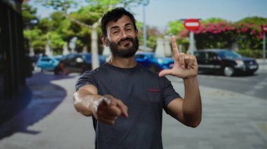 Sakallı İspanyol bir adam şakacı ezik hareketler yapıyor ve ağaçlar ve park edilmiş arabalarla dolu neşeli bir şehir caddesini işaret ediyor. Espri ve alaycılık yapıyor..