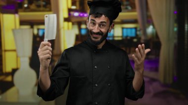 Şef üniforması giyen sakallı bir adam bir otelde satırı içeride tutuyor. Profesyonel bir mutfak ortamında İspanyol mirasına odaklanmış bir mutfak aktivitesi öneriyor..