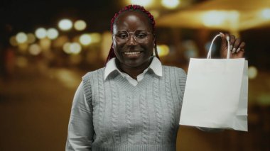 Gece şehir sokağında elinde alışveriş çantası olan bir kadın arka planda ışıklarla açık şehir manzarasını neşeli bir ifade ve moda tarzıyla gösteriyor..