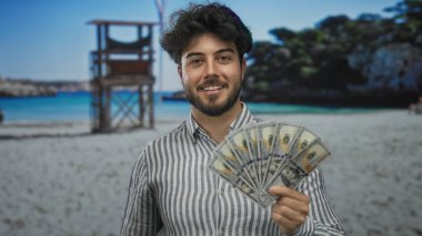 Bize dolar tutan sakallı genç adam sahilde gülümsüyor. Arka planda cankurtaran kulesi olan güneşli bir sahilde..