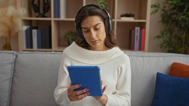 Kanepede dinlenen bir kadın rahat oturma odasında kulaklık takıyor ya da arka planda kitaplarla ders çalışıyor.
