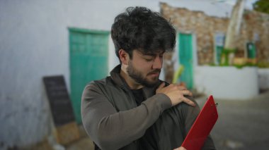 Güneşli bir günde İspanyol bir adam kırsal bir zemine karşı elinde bir panoyla omzunu tutarak rahatsızlık ya da yorgunluk hissi veriyor..