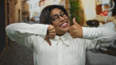 Gözlüklü, gülümseyen genç Latin kadın şehir sokaklarında başparmak kaldırma ve baş parmak kaldırma hareketleri yapıyor, şehir arka planında büyük boy güven gösterisi yapıyor..