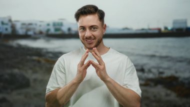 Bulutlu bir günde arka planda bulanık sahil binaları olan beyaz gömlekli genç adam deniz kenarında gülümsüyor..