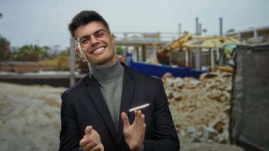 İspanyol bir adam açık hava şantiyesinde el çırpıyor, takım elbise ve gözlükle kendine güven ve tarz yayıyor..