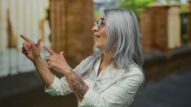 Kırlaşmış saçlı, dövmeli ve gözlüklü bir kadın arka planda bulanık marketlerle dolu açık bir sokakta hevesle el kol hareketi yapıyor..