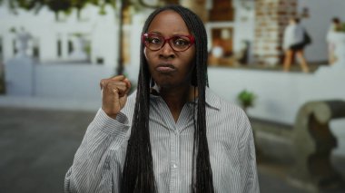 Şehir sokaklarında azim gösteren bir kadın, sıkılmış yumruğuyla güçlü bir poz veriyor ve kırmızı gözlüğü ve örgülü saçlarıyla ciddi bir ifade sergiliyor..