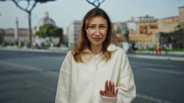 Beyaz kazaklı, belirsiz bir ifadeyle genç bir kadın, klasik mimari ve hafif ışıklandırma ile çevrili bir şehir caddesinde duruyor..