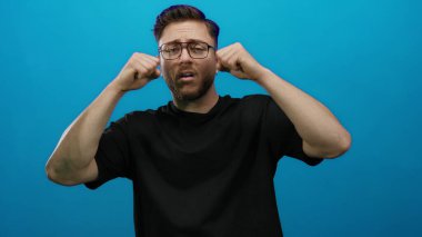 Sakallı genç bir adam, mavi bir arka plana karşı ağlayarak hareket ediyor. Duygularını yüzlerine yakın ifade ediyor. Siyah bir gömlek giyiyor..