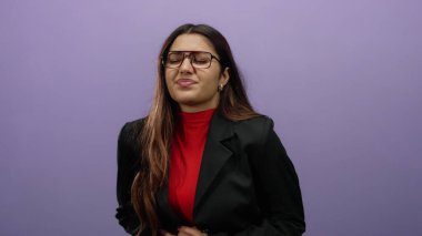 Gözlüklü ve siyah ceketli genç İspanyol kadın midesini mor bir duvara dayayıp rahatsızlık ya da mide ağrısına işaret ediyor..