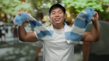 Şehrin bir caddesinde mavi beyaz bir eşarp takarak coşkuyla kutlama yapan genç adam, bulanık ağaçlar ve etrafındaki insanlarla şehir ortamında mutluluğu ifade ediyor..