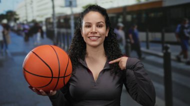 Basketbolu elinde tutan kadın, arka planda insanların dinamik açık hava ortamını ve spor atmosferini yakaladığı kentin hareketli sokaklarına güvenle tutunuyor..