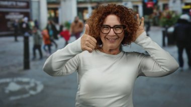 Middle-aged brunette hispanic woman on city street gives thumbs-up with people in background.