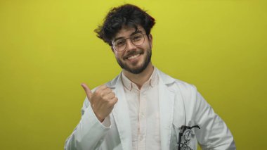 Beyaz önlüklü, sakallı ve gözlüklü genç İspanyol adam, sağlıklı bir ortamda onay ve pozitiflik öneren canlı sarı arka plana karşı emin adımlarla işaret ediyor..