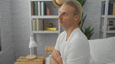 Orta yaşlı, beyaz, sarışın bir adam modern yatak odasının içinde, yatağında oturuyor, düşünceli ve rahat görünüyor..