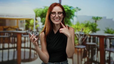 Şehir merkezindeki kafede terasta akıllı telefonuyla gülümseyen bir kadın, şehir manzarasının ortasında kızıl saçlı güzel ve hareketli sokak titreşimleri sergiliyor.