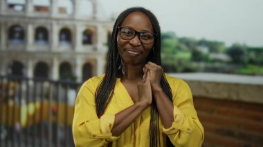 Roma 'da kolezyumun yanında gülümseyen bir kadın, gözlük takıyor ve sarı kıyafetler giyiyor, neşeli bir ifade sergiliyor ve uzun kıvırcık saçlı..