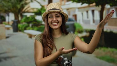 Hasır şapkalı İspanyol kadın turist, canlı şehir manzarasını elinde bir kamerayla sergiliyor, neşe dolu bir sokakta el kol geziyor..