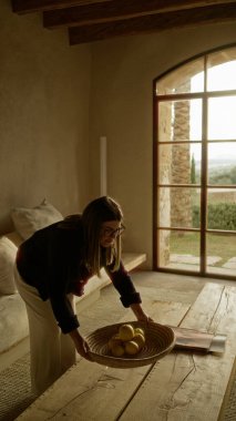 Akdeniz tarzı bir evde, ahşap kirişli ve taş duvarlı bir eve limon koyan bir kadın gözlük takıyor ve sıcak ve kırsal bir atmosfer yaratıyor..