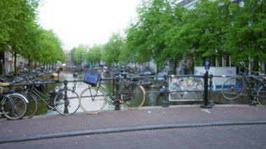 Amsterdam 'da, yeşil ağaçlar ve bulutlu bir gökyüzünün altında suyla kanalın yakınındaki bulanık bisikletler Hollanda' nın büyüleyici şehir manzarasını odaksız sokak manzarasıyla sergiliyor..