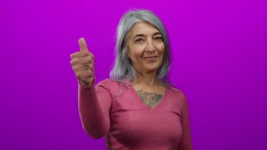 Gri saçlı yaşlı bir kadın pembe gömlekli, pembe renkli bir arka planda başparmak kaldırıyor. Stüdyo ortamında pozitifliğini ve onayını ifade ediyor..