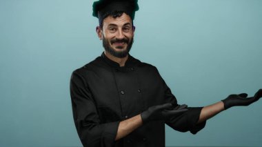 Siyah üniformalı İspanyol aşçı ve eldivenleri mavi arka plana karşı ellerini açmış mutfak bağlamında güvenle gülümsüyor..