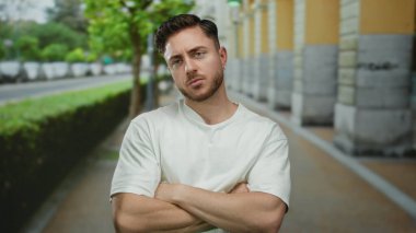 Sakallı ve beyaz gömlekli genç adam şehir caddesinde üstü kapalı duruyor. Bulanık binalar ve yeşil ağaçlar, kendine güveni ve günlük tarzını ifade ediyor..