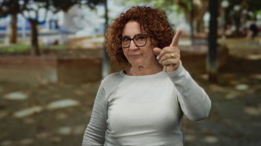 Woman gestures disapproval in park setting wearing glasses with brunette curly hair amid outdoor background suggesting a thoughtful moment during sunny day.