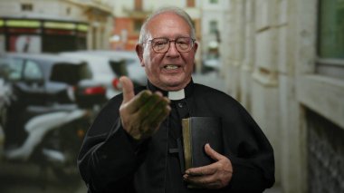 Başrahip gözlük takıyor kalabalık bir sokakta elinde bir kitapla dikiliyor. Arka planda arabalar ve binalar var..