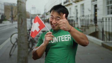 Şehir caddesinde elinde İsviçre bayrağı tutan genç adam yeşil gönüllü tişörtü giymiş açık hava kentsel manzarası gülümsüyor.