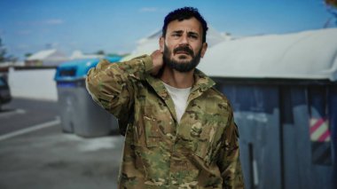 Askeri üniformalı İspanyol bir adam dışarıda çöp konteynırının yanında duruyor. Rahatsızlık içinde boynu ovalıyor, şehir ortamında stresi ya da yorgunluğu gösteriyor..