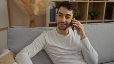Evdeki bir kanepede dinlenen genç adam, rahat bir oturma odasında, bitkiler ve kitaplarla telefon görüşmesi yaparak sıcak bir atmosfer yaratıyor..