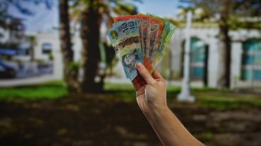 Canlı bir şehir parkında elinde Avustralya banknotları tutan bir adamın eli, kentsel bir ortamda finansal başarı ve para değişimi ruhunu yakalıyor..
