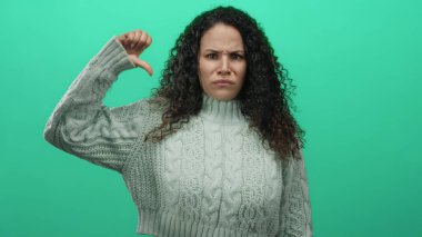 Süveter giyen genç İspanyol kadın, yeşil duvar arkaplanını işaret ediyor. Onaylamadığını belirtiyor ve yön gösteriyor..