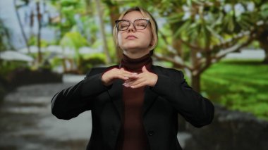 Parkta meditasyon yapan bir kadın yemyeşil bir alana kurulmuş, gözlük takmış, koyu renk ceketli, sakin ve sakin bir ortamda odaklanmış..