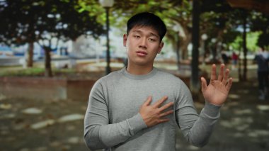 Genç Çinli bir adam, sakin bir açık hava parkında, gün ışığının altında el hareketleriyle duygularını ifade ederek iletişim için barışçıl ve çekici bir atmosfer yaratıyor..