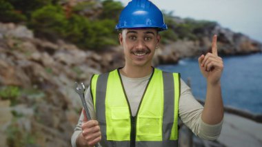 İngiliz anahtarı tutan düşünceli genç adam kumsalda deniz kenarında fikir jesti yapıyor, açık hava ortamına güveni yayıyor..