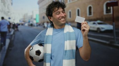 Sokak ortasında elinde futbol topu ve boş bir kartla gezen İspanyol bir adam. Gülümsemesi, canlı bir şehir sporu atmosferi olduğunu gösteriyor.