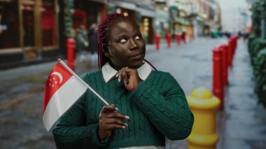 Kadın şehir caddesinde elinde Singapur bayrağı, yeşil kazak giyiyor ve gülümsüyor, şehir kültürünü ve çeşitliliğini gösteriyor..